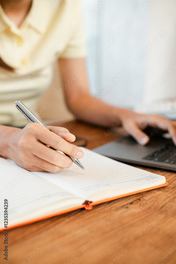 Poster close-up of a person writing in a notebook with a pen while using a laptop, showcasing productivity 