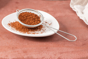 Fine cocoa powder in a metal sieve on a white plate, with some powder spilled around, set against a brown background