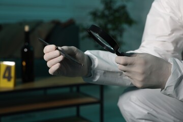 Forensic expert with tweezers examining hair through magnifying glass indoors, closeup