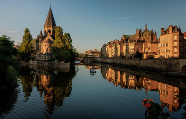 Obraz premium Historic riverside cityscape with a stone church, tree-lined banks, and reflective calm water illuminated by golden evening light.