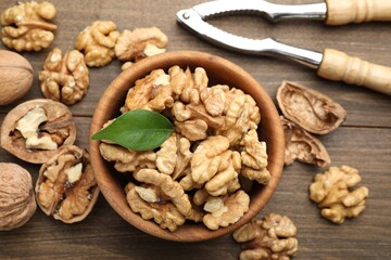 Peeled walnuts in bowl and nutcracker on wooden table, flat lay