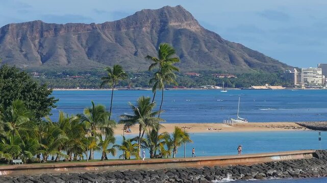 Aerial view of Waikiki Beach with Diamond Head crater, Honolulu skyline, blue ocean, jetty, gentle waves, and people walking along the jetty.