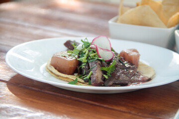 Close-up of a savory stewed beef tongue taco on a corn tortilla, topped with crispy potato chunks, radish slices, and fresh cilantro, served on a rustic wooden table.