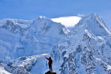 A trekker in a red jacket stands triumphantly on a rocky peak near Kanchenjunga Base Camp South, Nepal, surrounded by the majestic snow-covered Himalayas under a clear blue sky. © KhangRi Studio