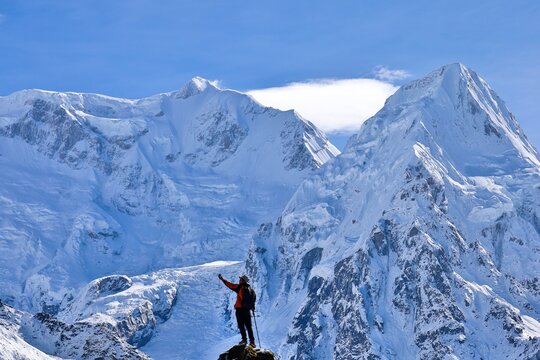 A trekker in a red jacket stands triumphantly on a rocky peak near Kanchenjunga Base Camp South, Nepal, surrounded by the majestic snow-covered Himalayas under a clear blue sky.