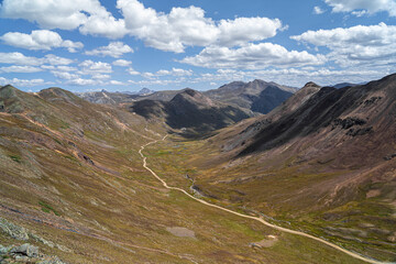 A stunning mountain valley featuring rugged ridges, deep valleys, and a meandering trail under a clear sky dotted with white clouds, evoking a sense of peace and adventure in nature.