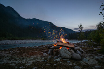 Campfire surrounded by stones with sparks flying into the twilight sky, situated by a tranquil river surrounded by mountains helping evoke peace and wilderness adventure.