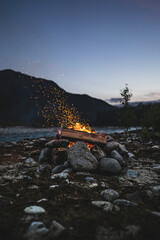 Vertical photo of a campfire surrounded by stones with sparks flying into the twilight sky, situated by a tranquil river surrounded by mountains helping evoke peace and wilderness adventure.