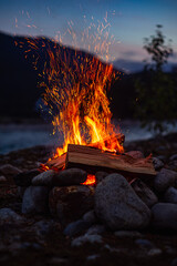 A close-up of a blazing campfire surrounded by stones, with sparks flying upwards, set in the backdrop of a peaceful outdoor evening scene under a twilight sky.