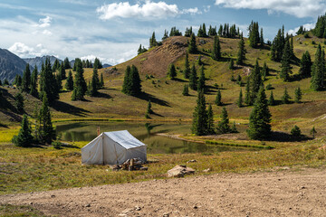A serene outdoor campsite featuring a white canvas tent near a calm lake surrounded by hilly greenery under a bright blue sky. Ideal for outdoor enthusiasts and nature exploration activities.