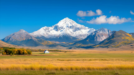 Fototapeta premium Snow-capped mountain peak rises above a golden autumn valley with a small white barn under a clear blue sky.