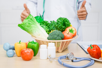 An Asian male nutritionist with a beard, wearing a lab coat, sits at a desk for an online consultation. His workspace includes fruits, vegetables, laptop, health supplements, promoting wellness.
