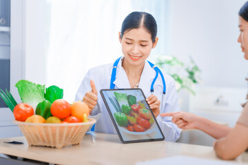 Young Asian female nutritionist in a white lab coat, working at a desk. She shows tablet with...