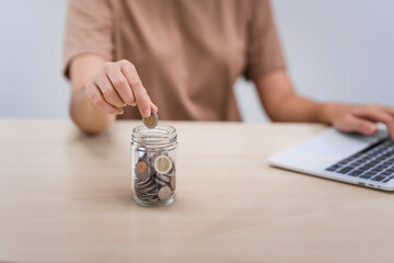 A young Asian woman in a light brown t-shirt, sitting with a piggy bank, coin, calculator, mobile, and pen. She focuses on financial stability, saving for emergencies, education, and future goals.
