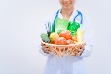 A young Asian woman nutritionist in a white lab coat, holding fruit and vegetable basket, smiling happily against white background. dietary nutrients, metabolism, and promoting health.