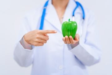 Young Asian nutritionist in a white lab coat, holding Green bell pepper and smiling happily against...