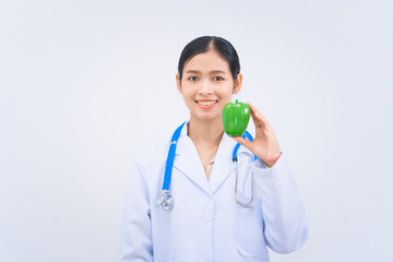 Young Asian nutritionist in a white lab coat, holding Green bell pepper and smiling happily against...