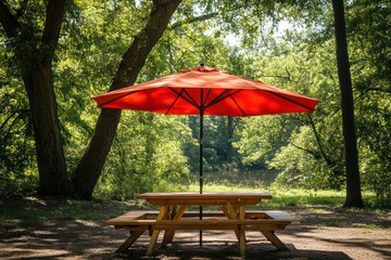 Obraz premium A picnic-style table with wooden benches and a bright red umbrella, surrounded by trees in a park.