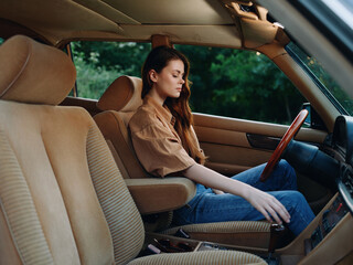 Beautiful young woman sitting in a vintage car, wearing casual attire, showcasing a serene expression, surrounded by a natural green background.