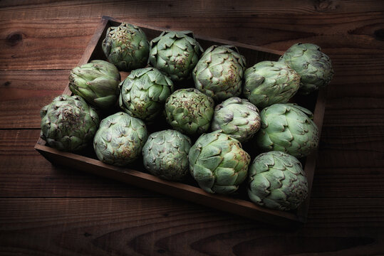A wood crate filled with fresh picked artichokes on a dark wood tavle with strong side light.