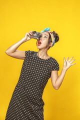 Surprised Asian girl dressed in pin-up style with vintage camera in her hands doing photo shoot on bright yellow background. Isolated.