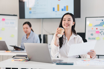 Fototapeta premium cheerful businesswoman holding coffee cup and paper, working in modern office with charts and laptops
