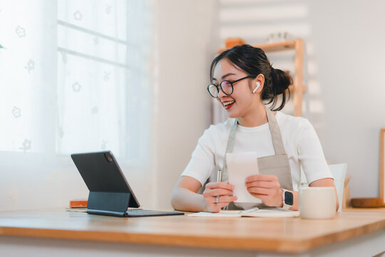 cheerful woman wearing glasses and apron works on tablet at wooden desk in bright home office - Powered by Adobe