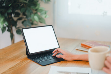 Fototapeta premium person working on tablet with keyboard case on wooden desk, holding coffee cup, surrounded by notebooks and plant