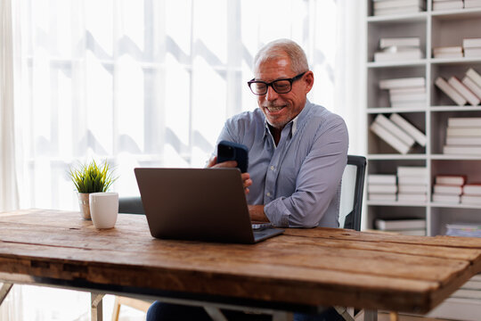 Smiling mature businessman holding smartphone sitting workplace office. Middle aged manager ceo using cell phone mobile apps and laptop. technology and solutions for business development