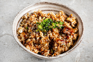 A delicious rice bowl featuring tender pieces of chicken, drizzled with a rich sauce and topped with sesame seeds and fresh green onions, set on a textured stone surface. 