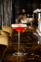 On a lavish marble table, a vibrantly colored cocktail awaits. The stylish drink is set in a delicate glass, capturing the ambiance of an upscale bar. Patrons mingle nearby. 