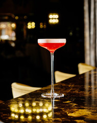 A beautifully crafted cocktail with a vibrant hue sits gracefully on a glossy table. The warm ambiance of the bar adds charm to the evening gathering, inviting patrons to enjoy.