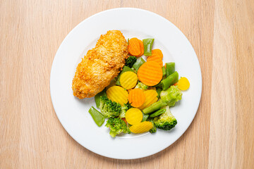 A golden-brown crispy chicken breast rests beside a vibrant array of steamed vegetables, including broccoli, peas, and sliced carrots. The dish is artfully arranged, showcasing freshness. 
