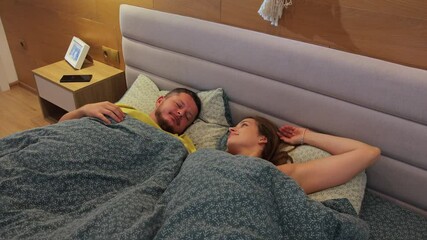 A young couple in bed wake up in the morning. They open their eyes and greet each other. The man takes the smartphone and looks at the information on the screen. His wife kisses his forehead. - Powered by Adobe