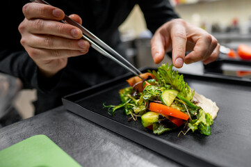 A chef skillfully arranges a vibrant salad on a black plate using tongs. Fresh greens, tomatoes, and vegetables highlight the artistry of culinary presentation in a modern kitchen.