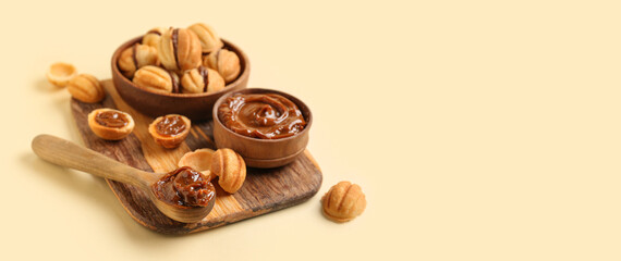 Wooden board and bowl of sweet walnut shaped cookies with boiled condensed milk on yellow background