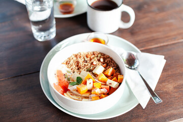 A healthy granola bowl with yogurt, mango, papaya, banana, honey, coconut flakes, and flax seeds. Served with a glass of water and a cup of coffee, creating a perfect balanced breakfast