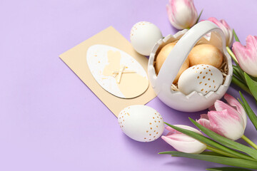 Basket with Easter eggs, tulips and festive postcard on lilac background