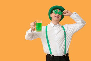 Happy young man in leprechaun's hat with party glasses and mug of beer on yellow background. St....