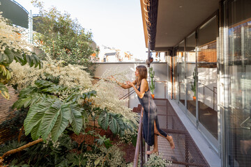 A woman in a minimalist dress leans on a railing, enjoying lush greenery on a modern balcony. The...