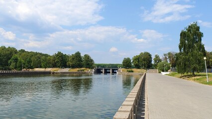 Fototapeta premium The banks of the river flowing through the city are concreted, with tile walkways and light poles along them. A dam blocks the river. Trees grow on the grassy lawns. Sunny summer weather and blue sky