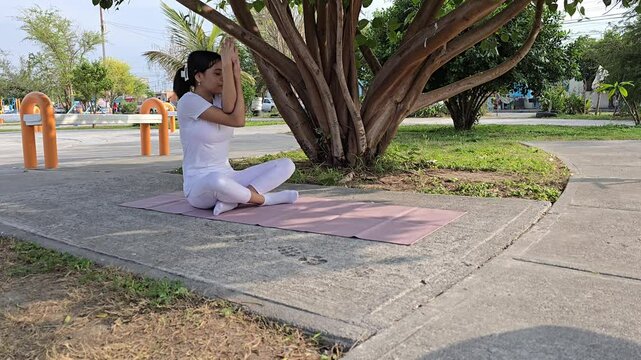 Latina teenager doing yoga under a tree at the park (Seated eagle pose)