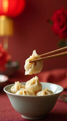 Creative holiday culinary concept: chopsticks with plate of stuffed dumplings with sauce, cup of tea and blurred red flowers on oriental lanterns background for Chinese New Year. Space for text.