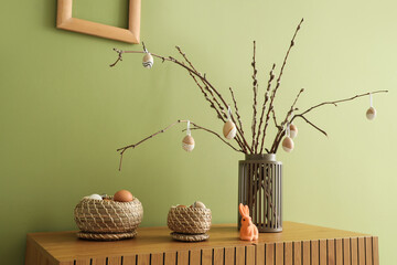 Vase with tree branches and Easter eggs on commode near green wall