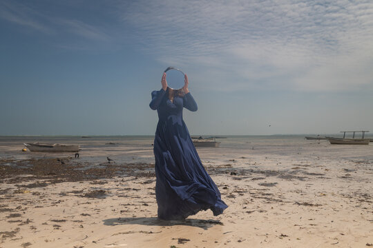 art portrait of woman in long blue dress on the beach holding round mirror hiding her face