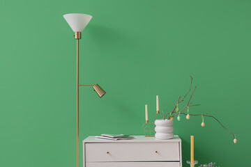 Vase with tree branches, Easter eggs and candles on commode near green wall in room