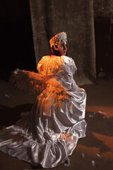 Beautiful young brunette dressed as an angel kneels in a dimly lit room, draped in an ornate white...