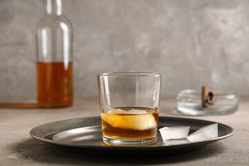 Glass of cold rum with ice cubes on plate on table against grey background. Closeup