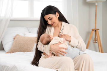 Young woman in pajamas breastfeeding her baby while sitting on bed at home
