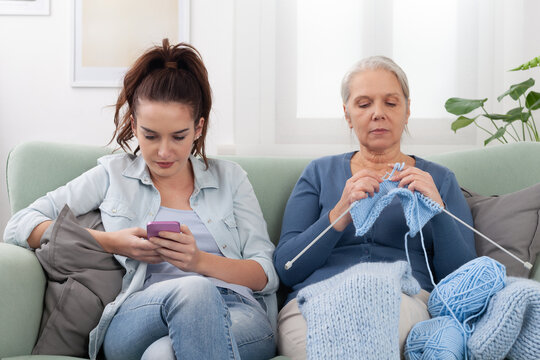 At home on the couch, a Millennials daughter uses her smartphone to browsing while her senior mother Boomer sits nearby knitting. Contrast of two generations of different lifestyles, generation gap